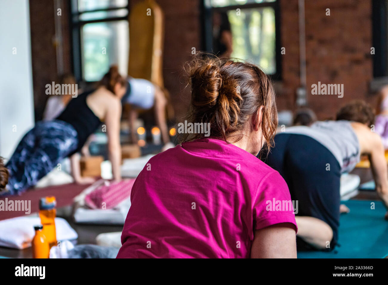 A closeup and rear view of a lady wearing a pink shirt with brunette hair tied back during a calming yogic class practicing a traditional routine of postures. Stock Photo