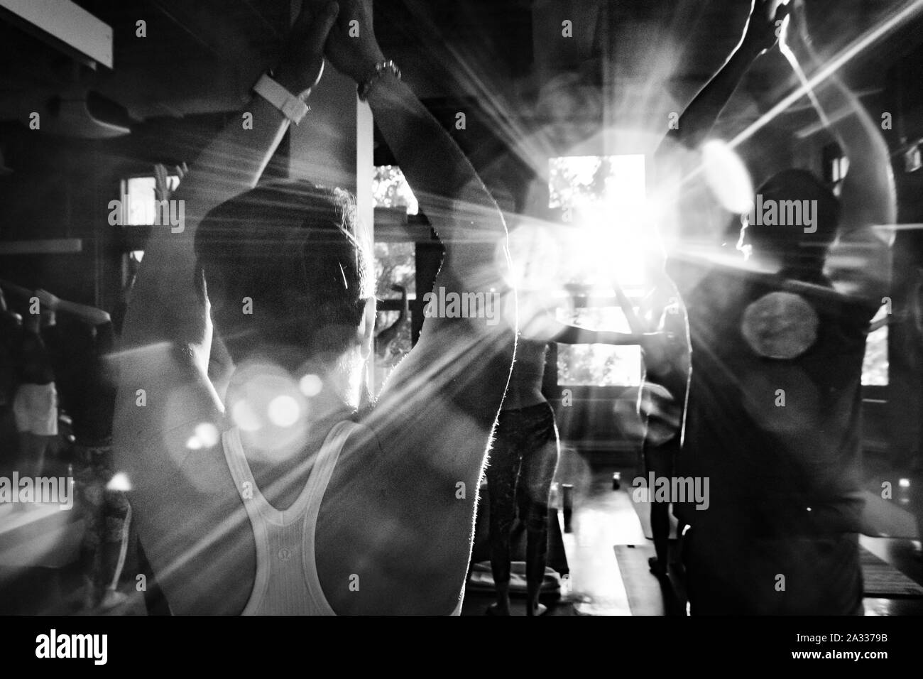 A monochrome view of people practicing 108 surya namaskar movements during a workshop. Class face the natural light glaring through windows as they salute sun. Stock Photo