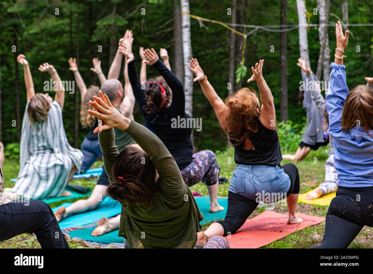 A group of people of all age groups are seen in warrior I pose (virabhadrasana I), during an outdoor yoga session as part of a multicultural celebration. Stock Photo
