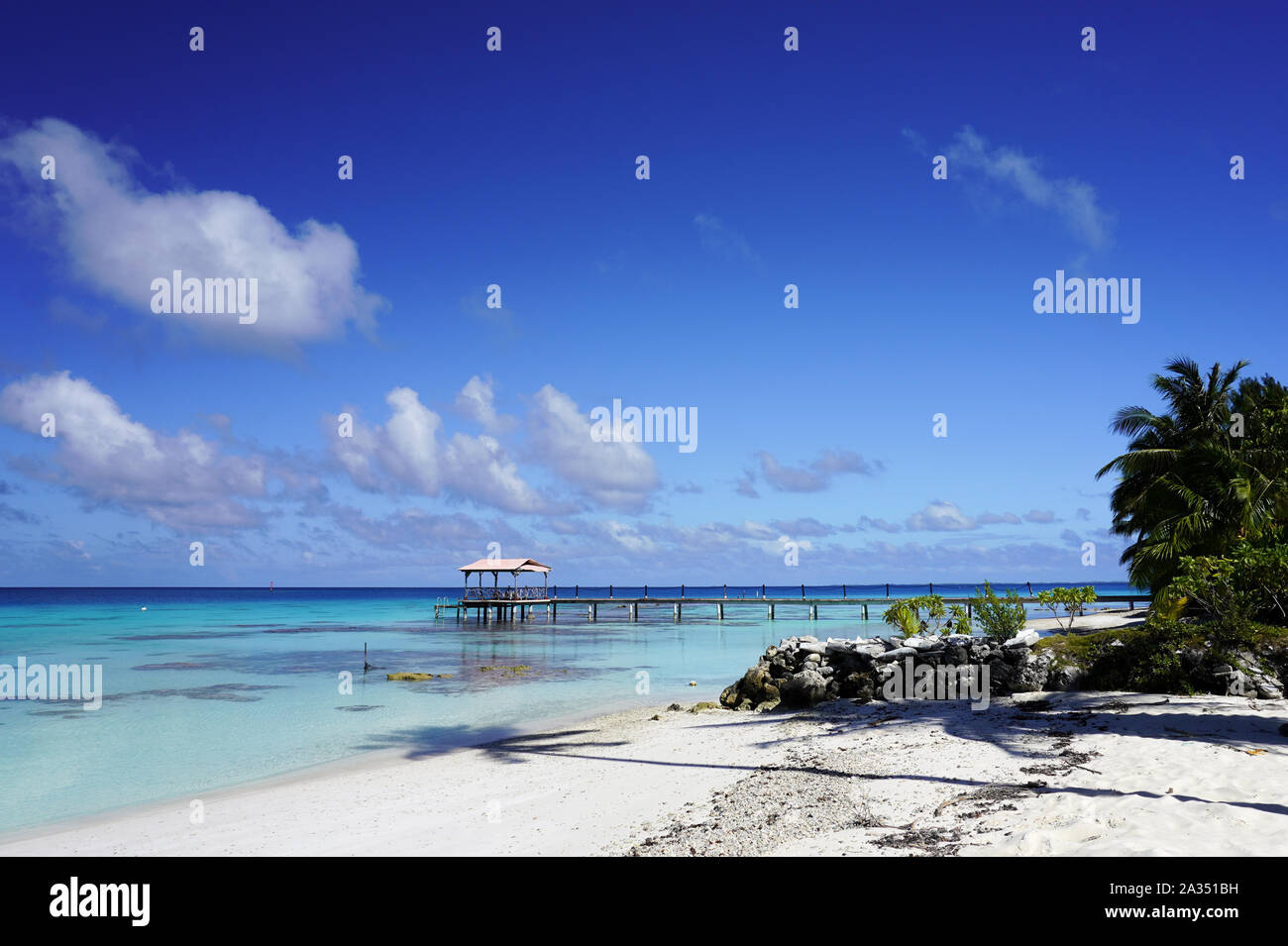 View of a dock from a sandy beach with palm trees leading into a blue lagoon on a tropical island Stock Photo