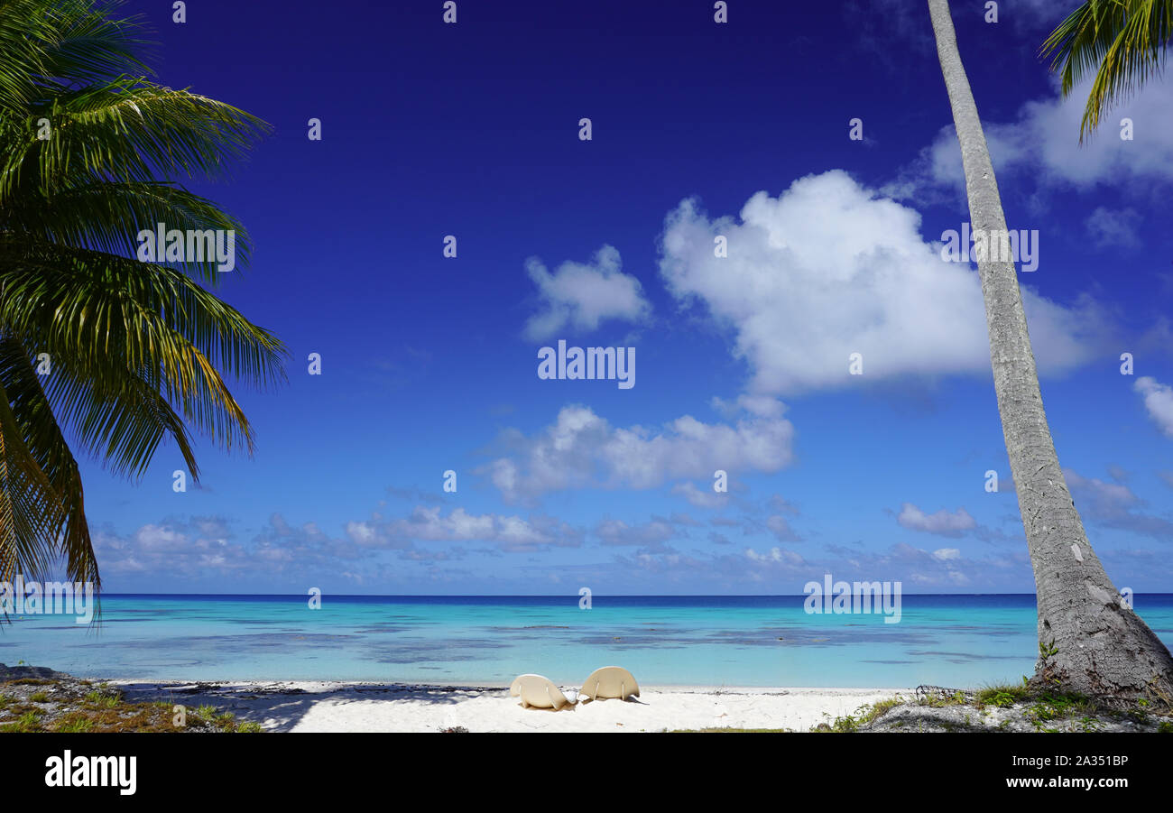 Two chairs on a sandy beach with palm trees and a turquoise lagoon under a bright blue sky with clouds on the island of Fakarava in French Polynesia Stock Photo
