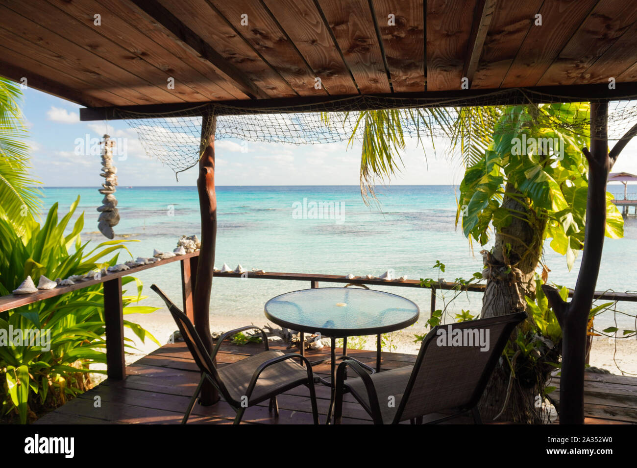 View of a blue tropical lagoon from the wooden deck of a bungalow with a table and chairs and framed by palm trees and seashells Stock Photo