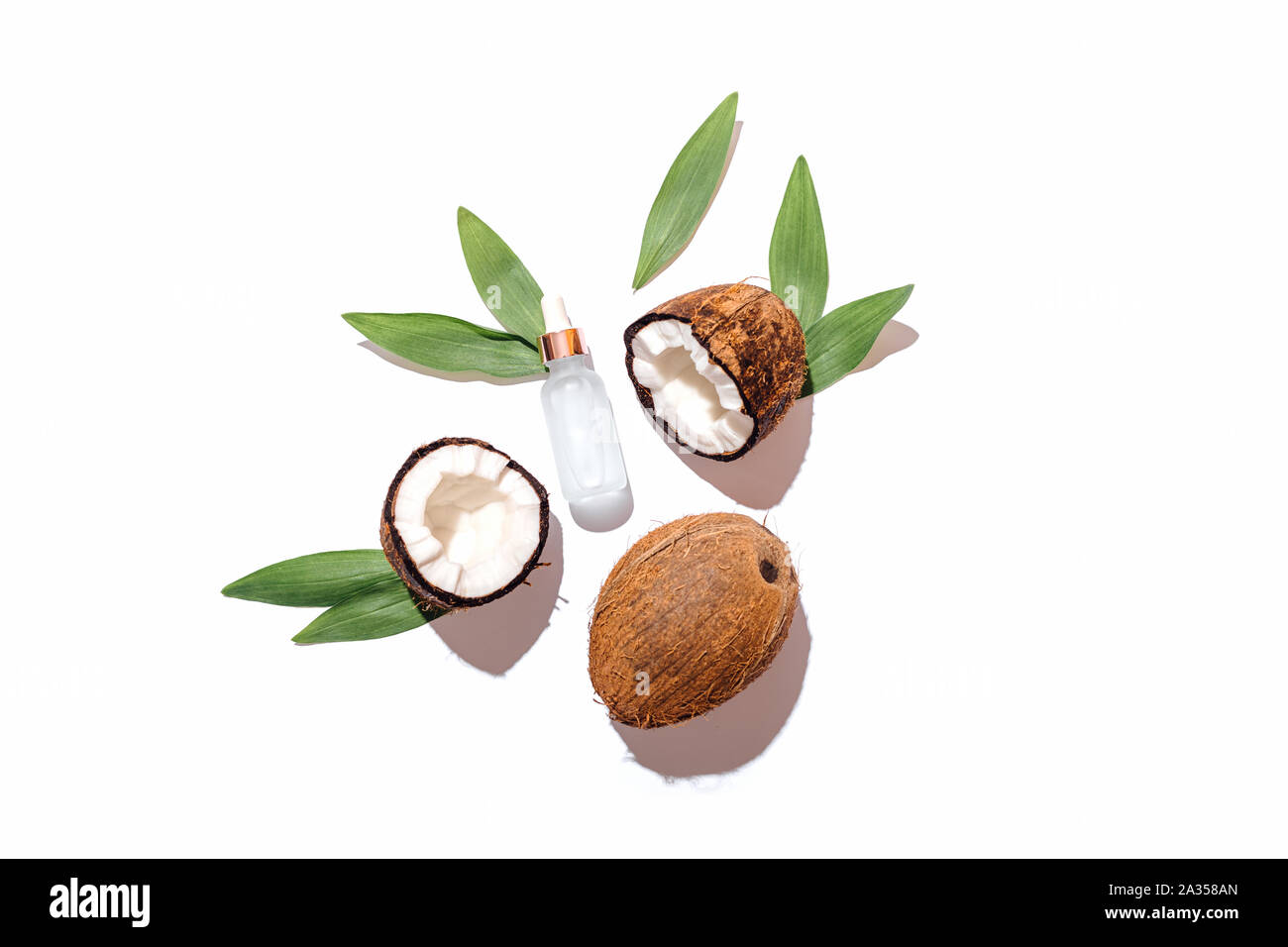 Fresh ripe coconuts with green leaves next to bottle of cosmetic serum on white background, flat lay composition. Stock Photo