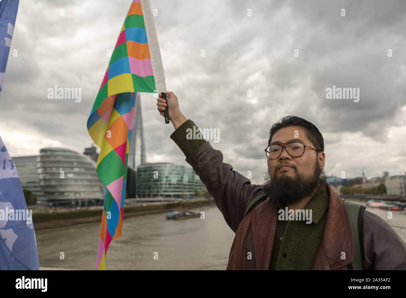 London, UK. 5th Oct, 2019. Chinese artist Badiucao with the Lennon Wall flag on Tower Bridge.  Chinese artist Badiucao and supporters join hands in a human chain, a showing of solidarity with protesters in Hong Kong. The Lennon Wall flag, symbolising Hong Kongs fight for freedom is also symbolically raised. Penelope Barritt/Alamy Live News Stock Photo