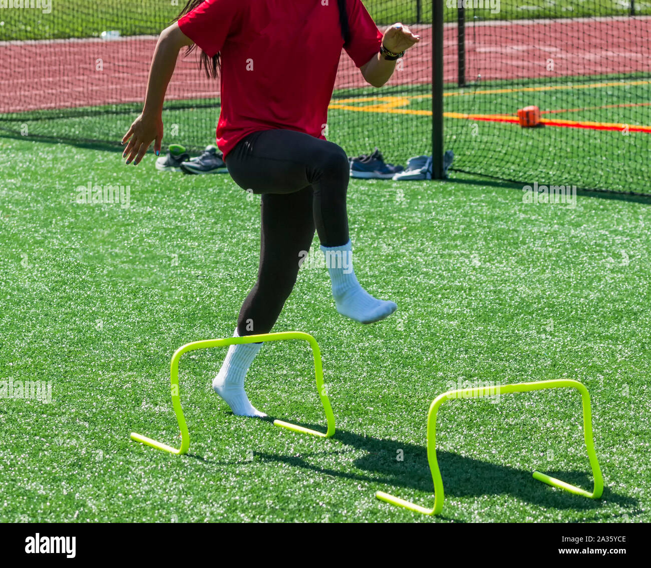 A high school female track and field athlete is performing form drills with no shoes on by steping over yellow mini hurdles on a green turf field. Stock Photo