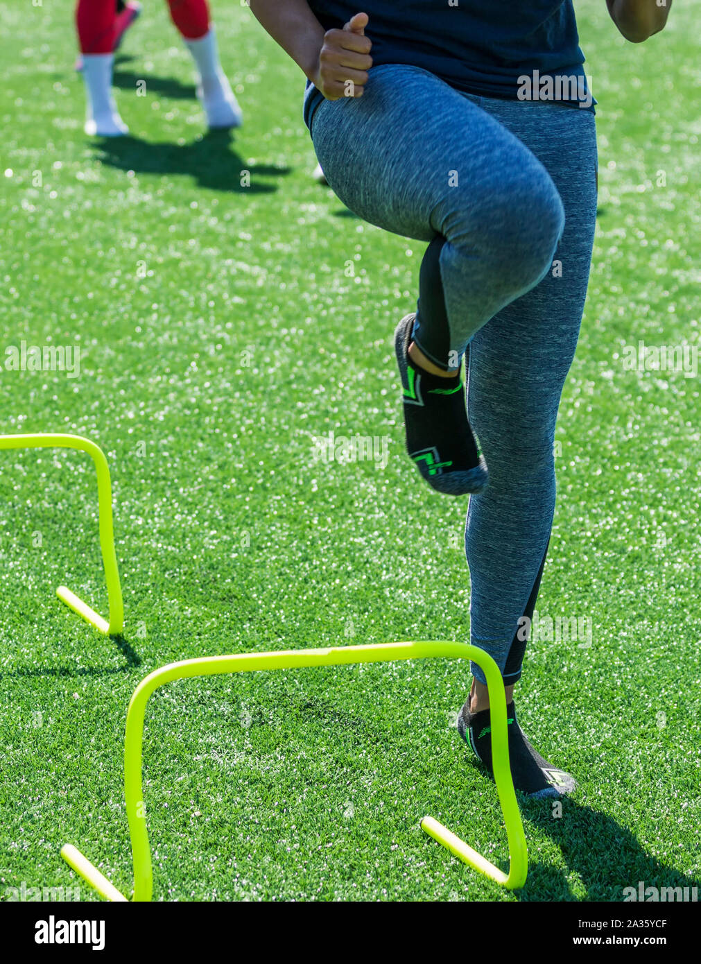 A high school female athlete performs running drills over mini hurdles while wearing socks but no shoes on a green turf field. Stock Photo