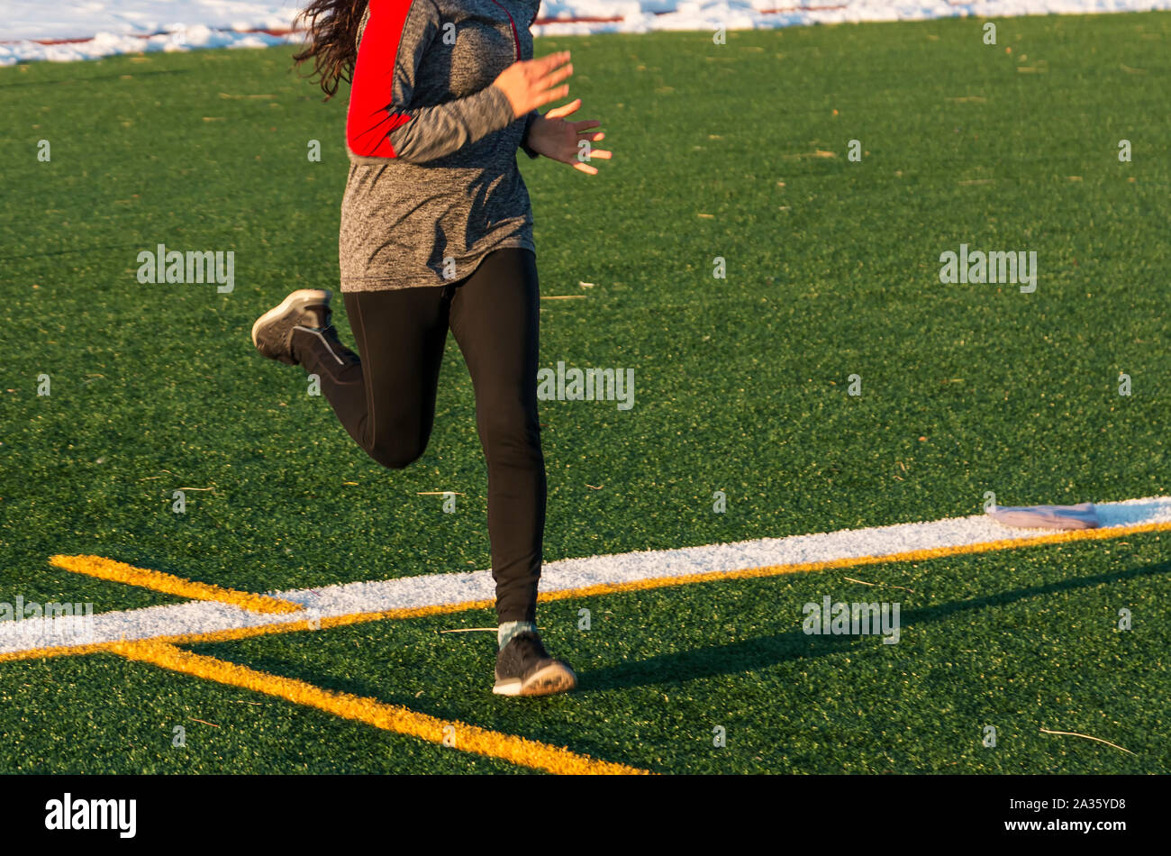 A high school female runner is running fast on a green turf field iin the winter, with snow lining the track behind her. Stock Photo