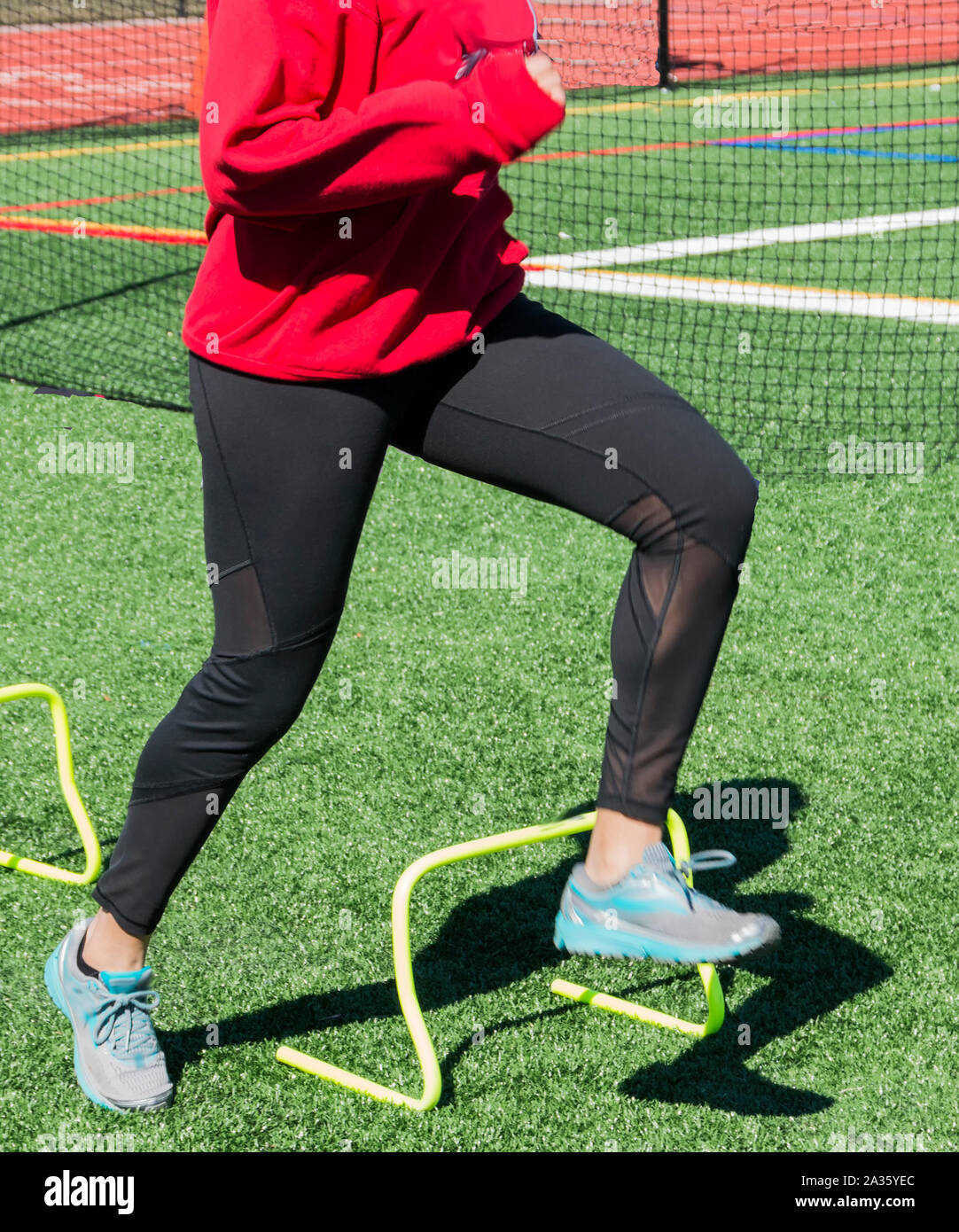 A high school athlete is stepping, running, over yellow mini hurdles during strength and speed training practice on a green turf field. Stock Photo