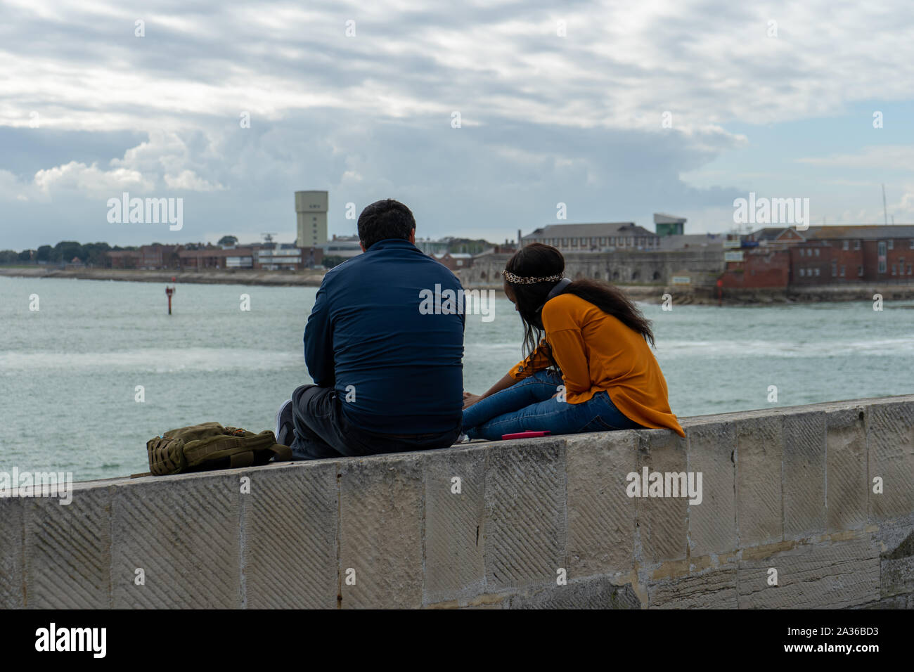 A multicultural couple sitting on a wall at the seaside chatting while looking out to sea at the view Stock Photo