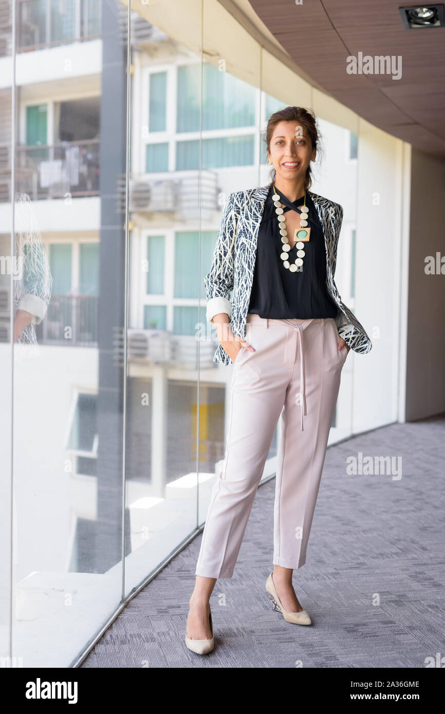 Full body shot of happy Hispanic businesswoman smiling by the window at office building Stock Photo