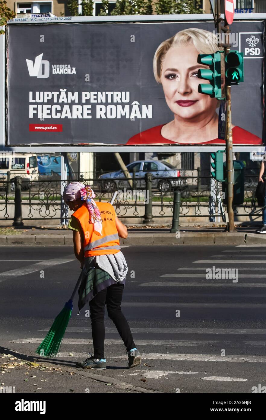 Bucharest, Romania - October 01, 2019: A woman sanitation worker sweeps a street in front of a presidential campaign billboard  of Viorica Dancila, th Stock Photo