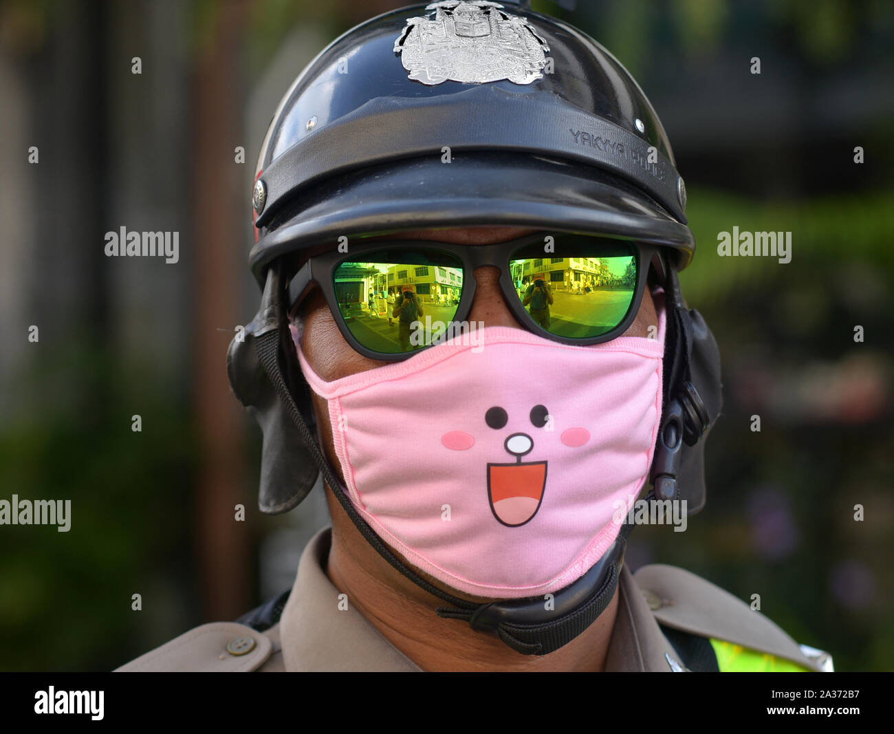 An unidentifiable Royal Thai motorcycle police officer wears mirrored sunglasses and a funny protective face mask in pink. Stock Photo