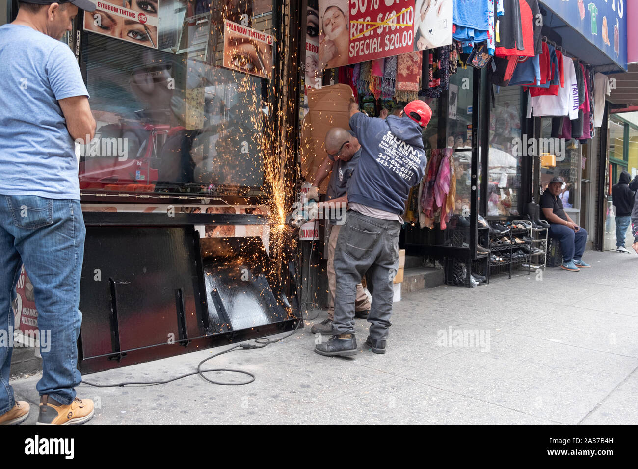 A structural iron & steel worker works on a storefront door while a colleague shields the adjacent store from sparks. In Jackson Heights, Queens, NYC. Stock Photo