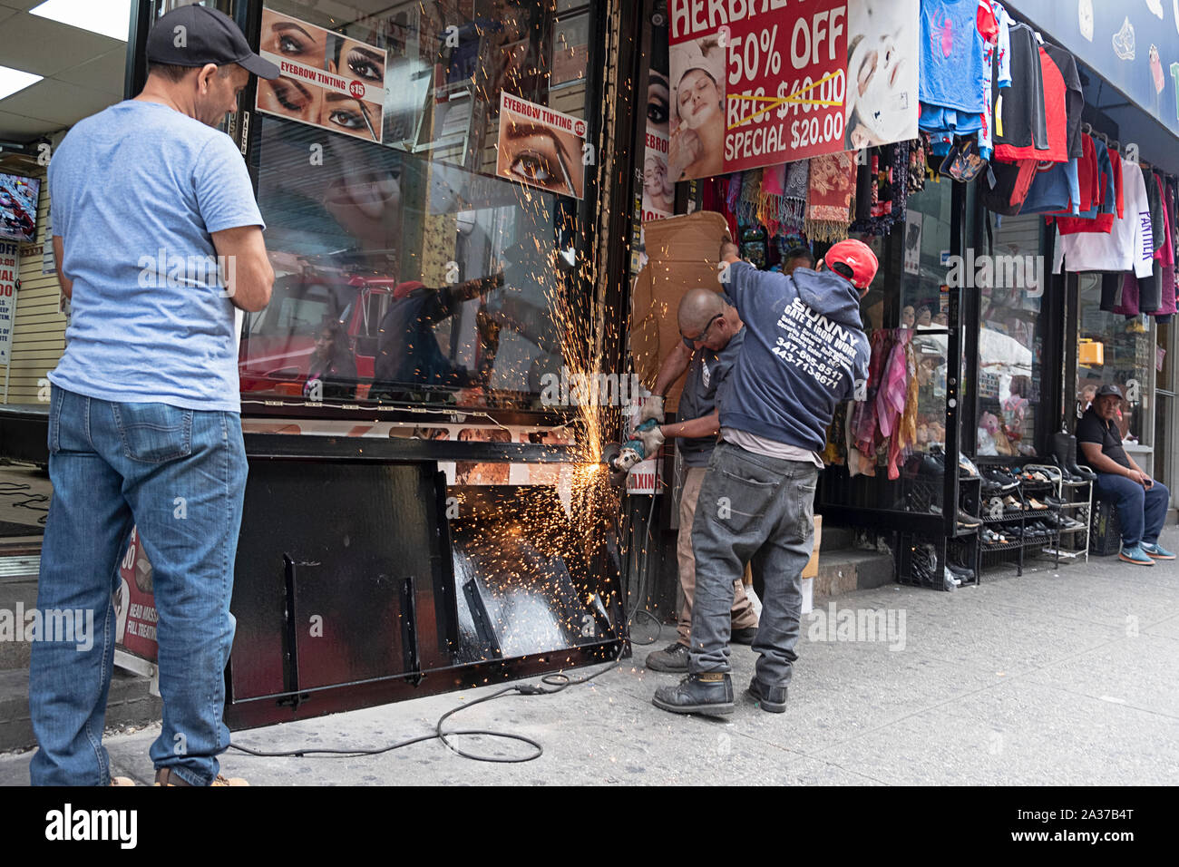 A structural iron & steel worker works on a storefront door while a colleague shields the adjacent store from sparks. In Jackson Heights, Queens, NYC. Stock Photo