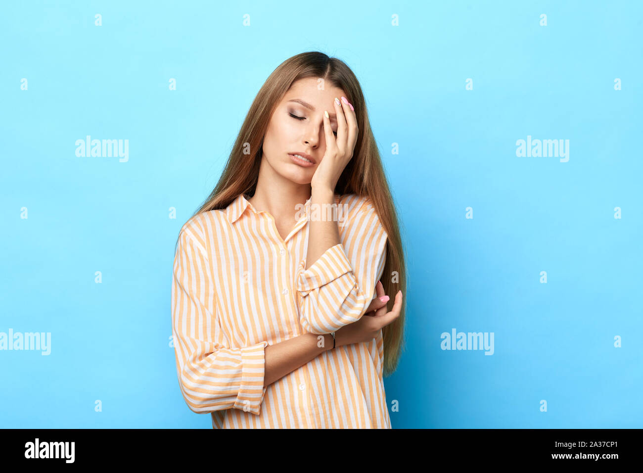 Beautiful exhausted woman feels sleepy, covers face with palm, sighs from tiredness, wears fashion shirt, isolated on blue background. Negative feelin Stock Photo