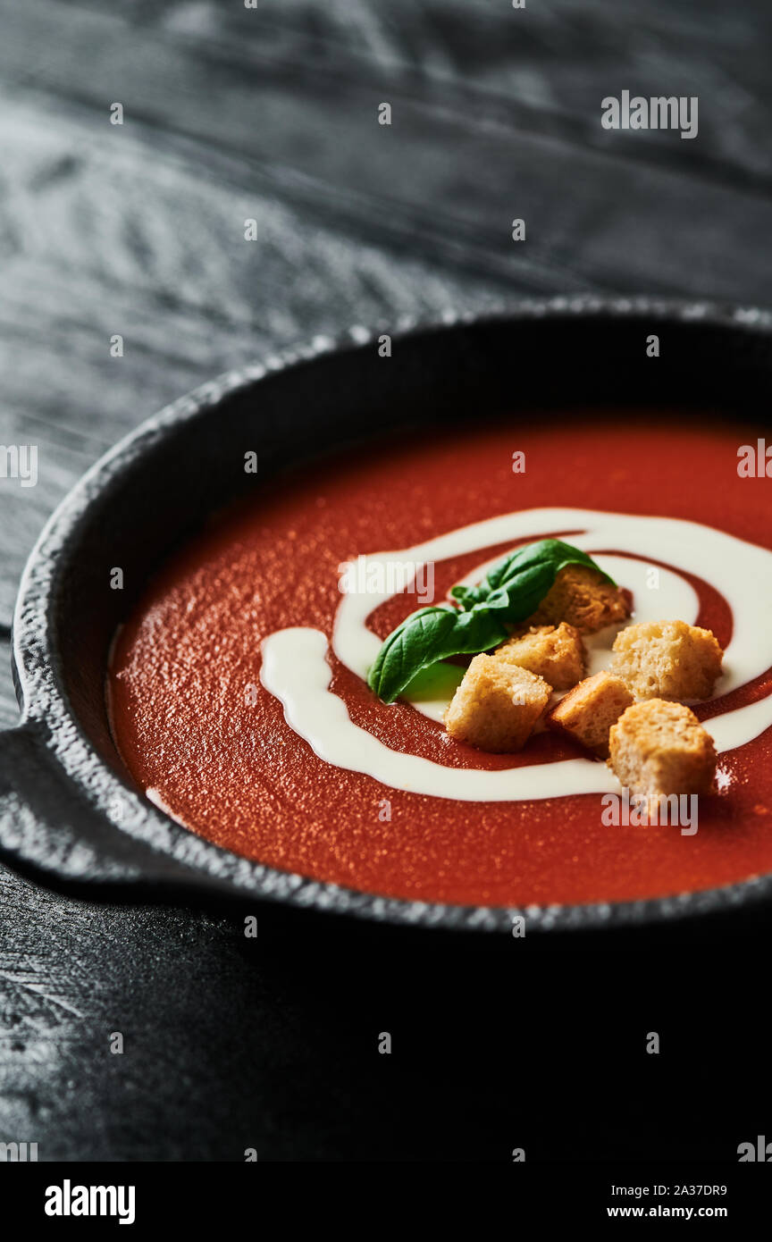 Delicious tomato soup with cream, croutons and basil in a black bowl on black wooden background. Copy space for text. Selective focus. Stock Photo