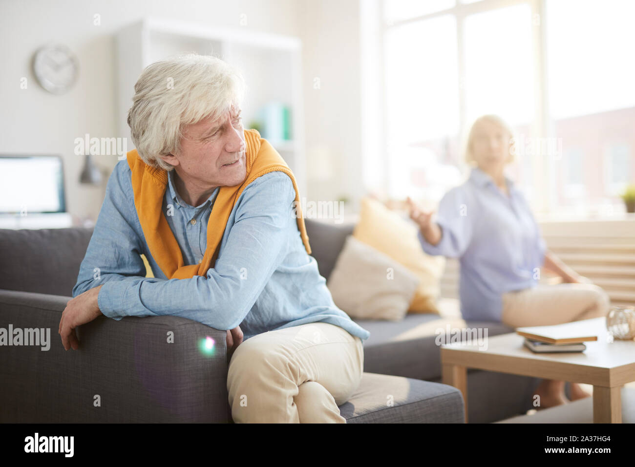 Portrait of senior couple fighting sitting on opposite sides of sofa at home, focus on angry husband in foreground, copy space Stock Photo