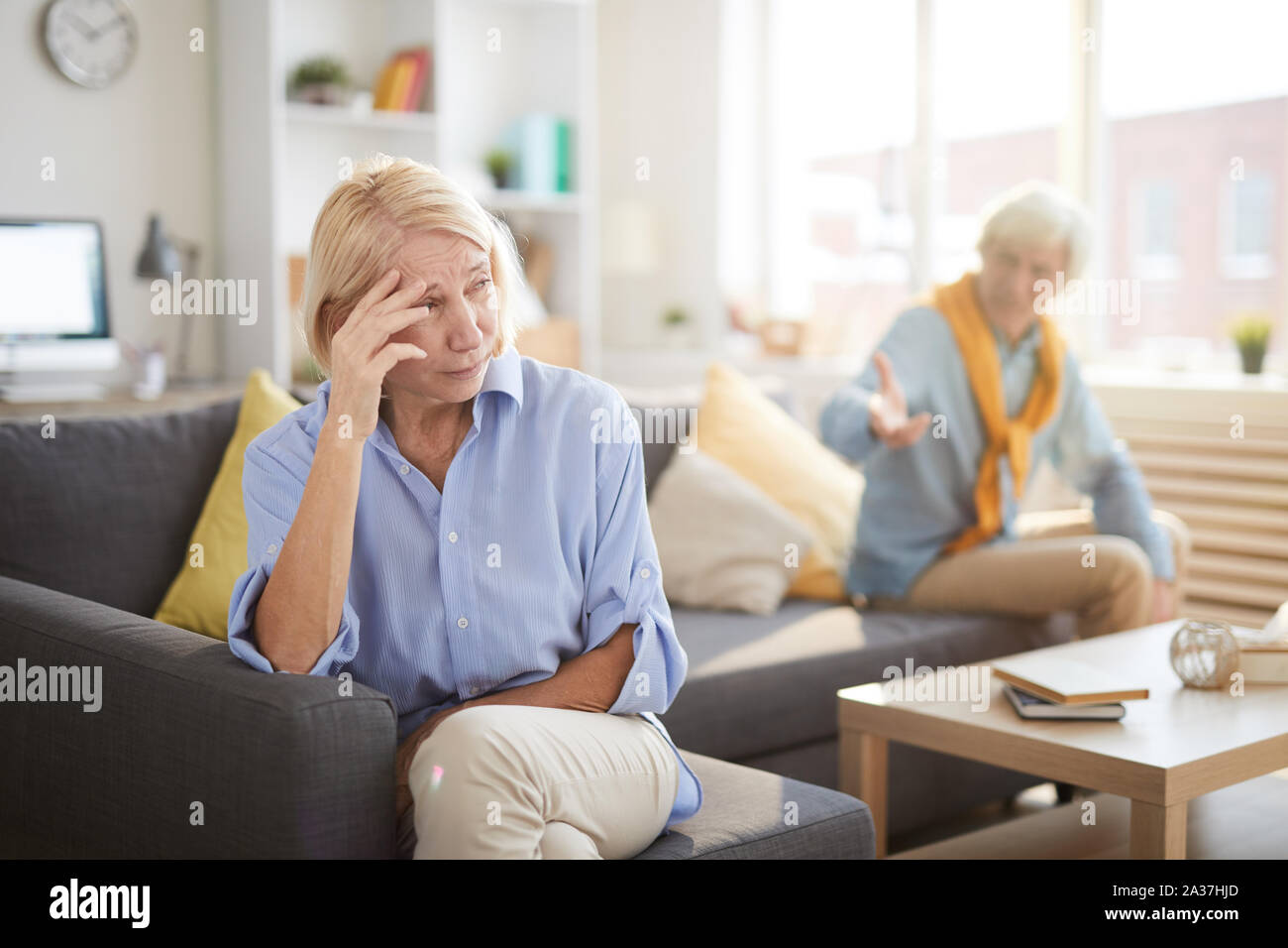 Portrait of senior couple fighting sitting on opposite sides of sofa at home, focus on crying wife in foreground, copy space Stock Photo