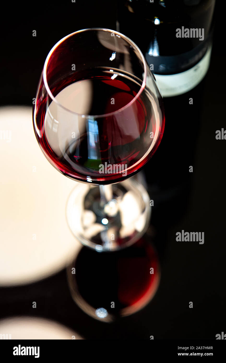 a glass of red wine in top view (focus on margin) with a blurry bottle of wine and ablack background with  white light reflection Stock Photo
