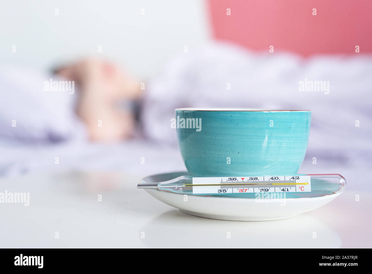 Cup of tea and thermometer with high temperature with a flu sick woman in bed on the background. Stock Photo