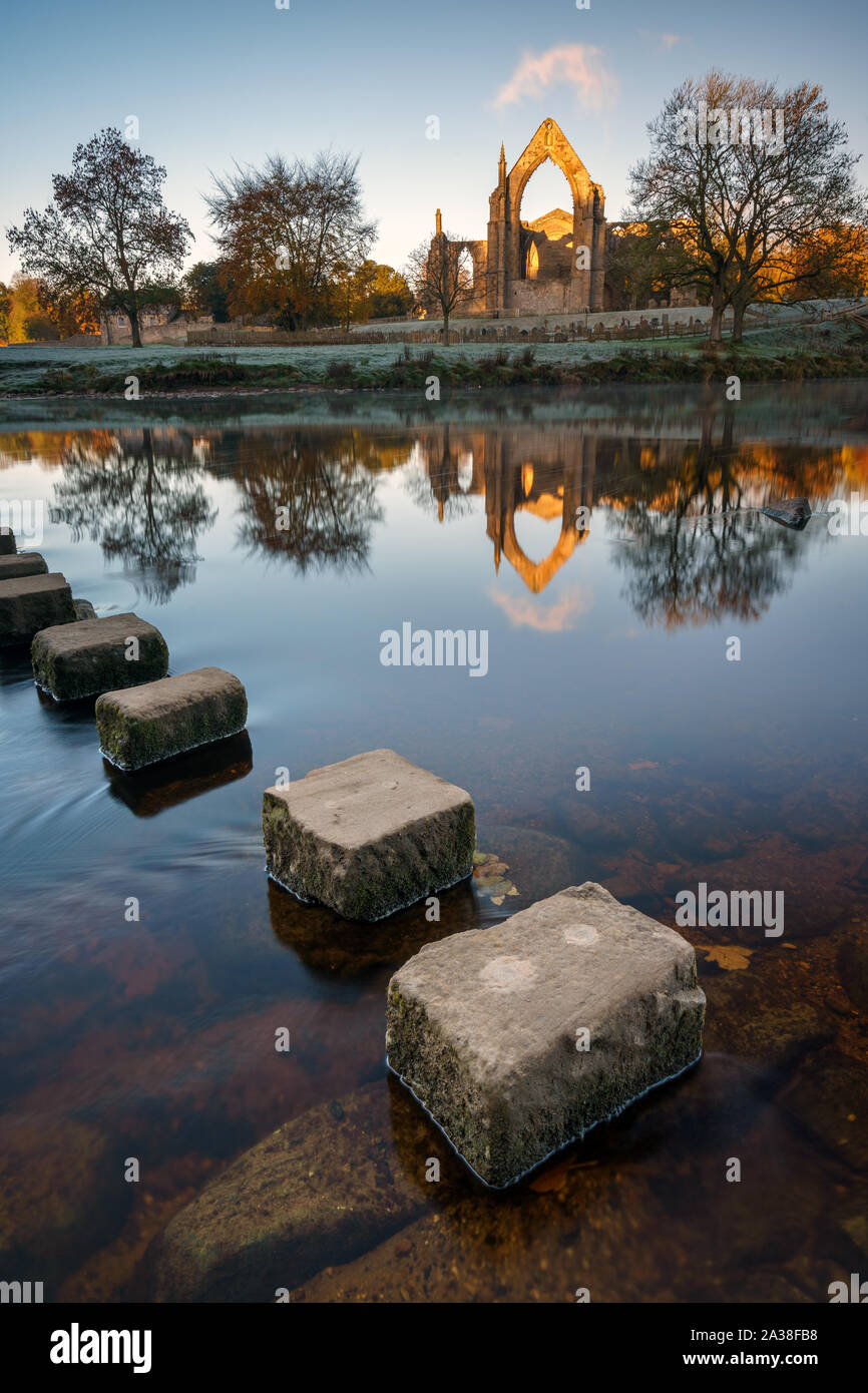 The ruins of Bolton Abbey catch the first direct light on a frosty autumn morning with stepping stones across the River Wharfe in the foreground. Stock Photo