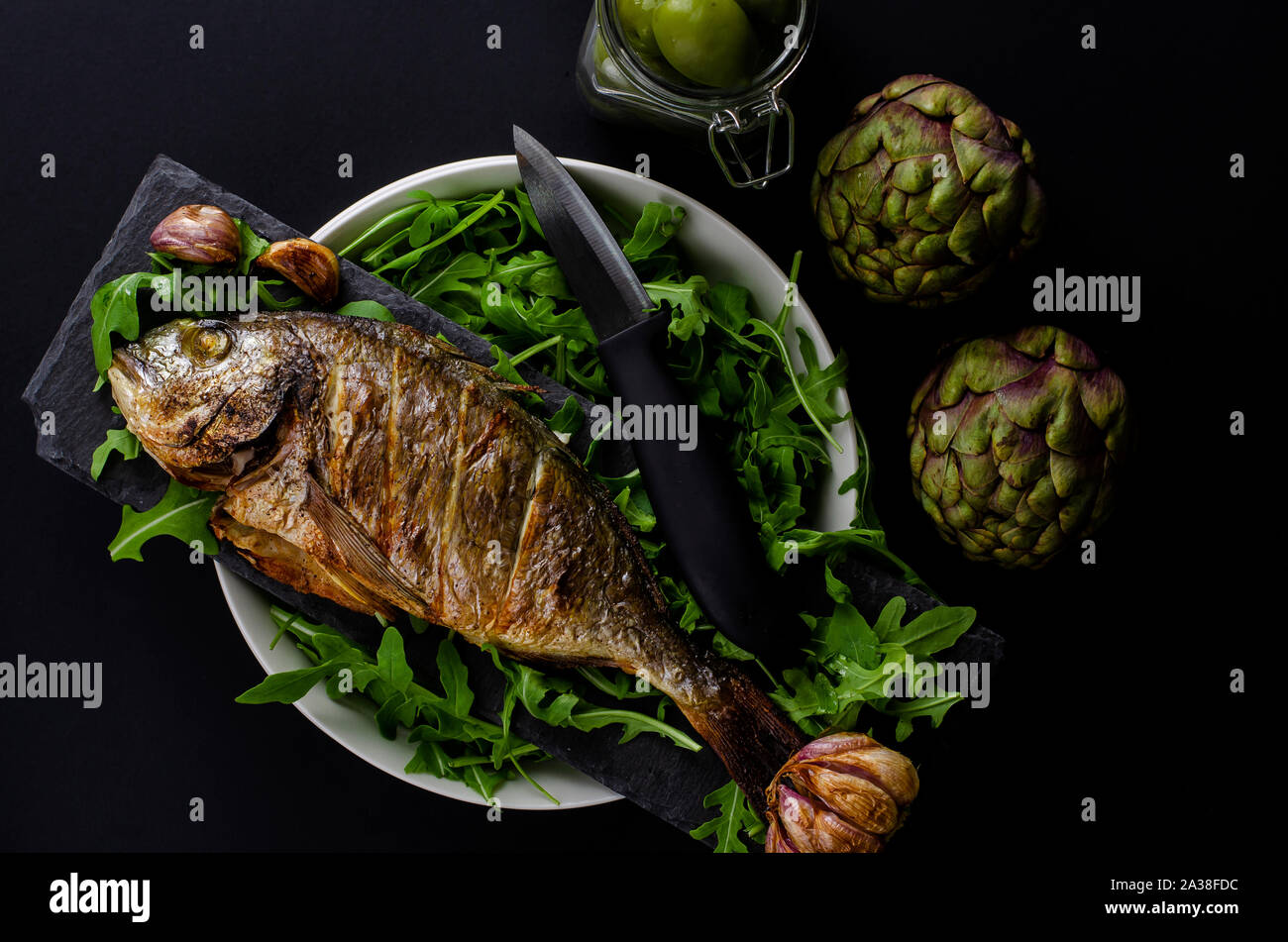 Top view of baked sea bream on a bowl with arugula, artichokes and knife on black background. Mediterranean food concept. Stock Photo