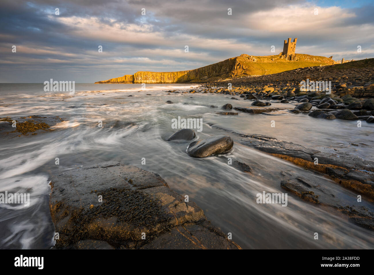 The ruins of Dunstanburgh Castle perch on the cliff above Embleton Bay as the North Sea waves break around the dolerite boulders along the shoreline. Stock Photo