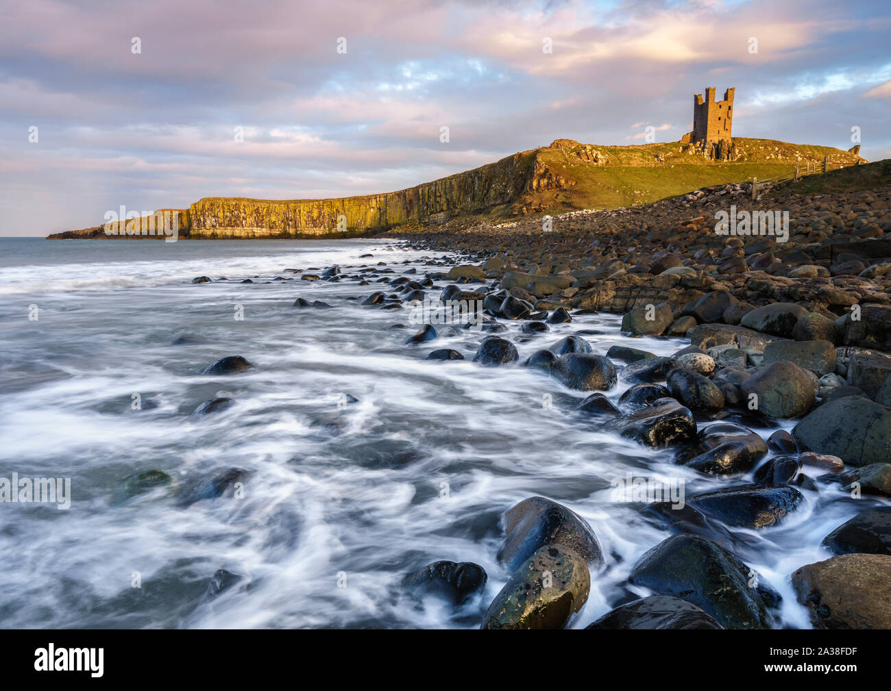 The ruins of Dunstanburgh Castle perch on the cliff above Embleton Bay as the North Sea waves break around the dolerite boulders along the shoreline. Stock Photo