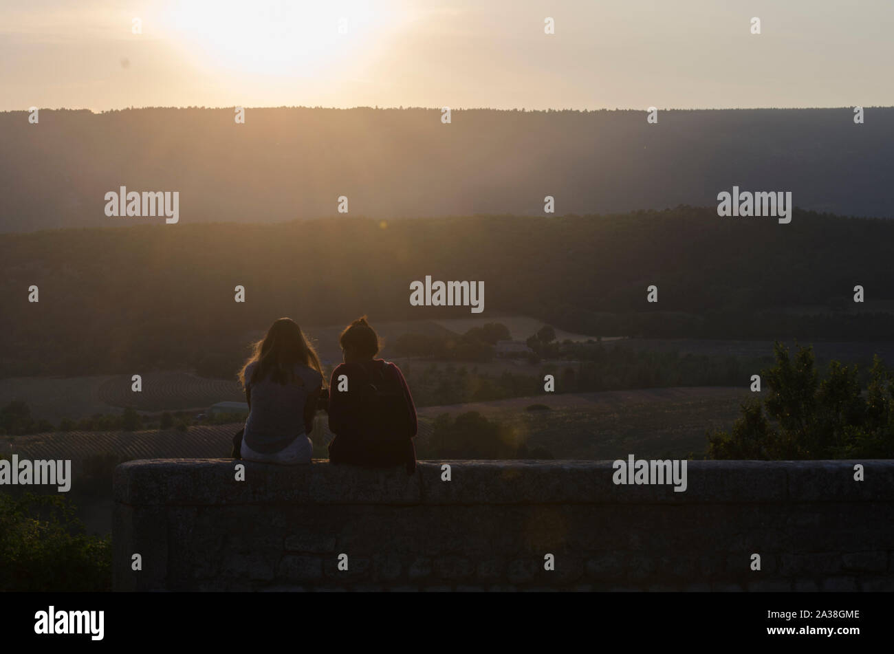 Rear view of two girls sitting on a wall looking at the sunset, France Stock Photo