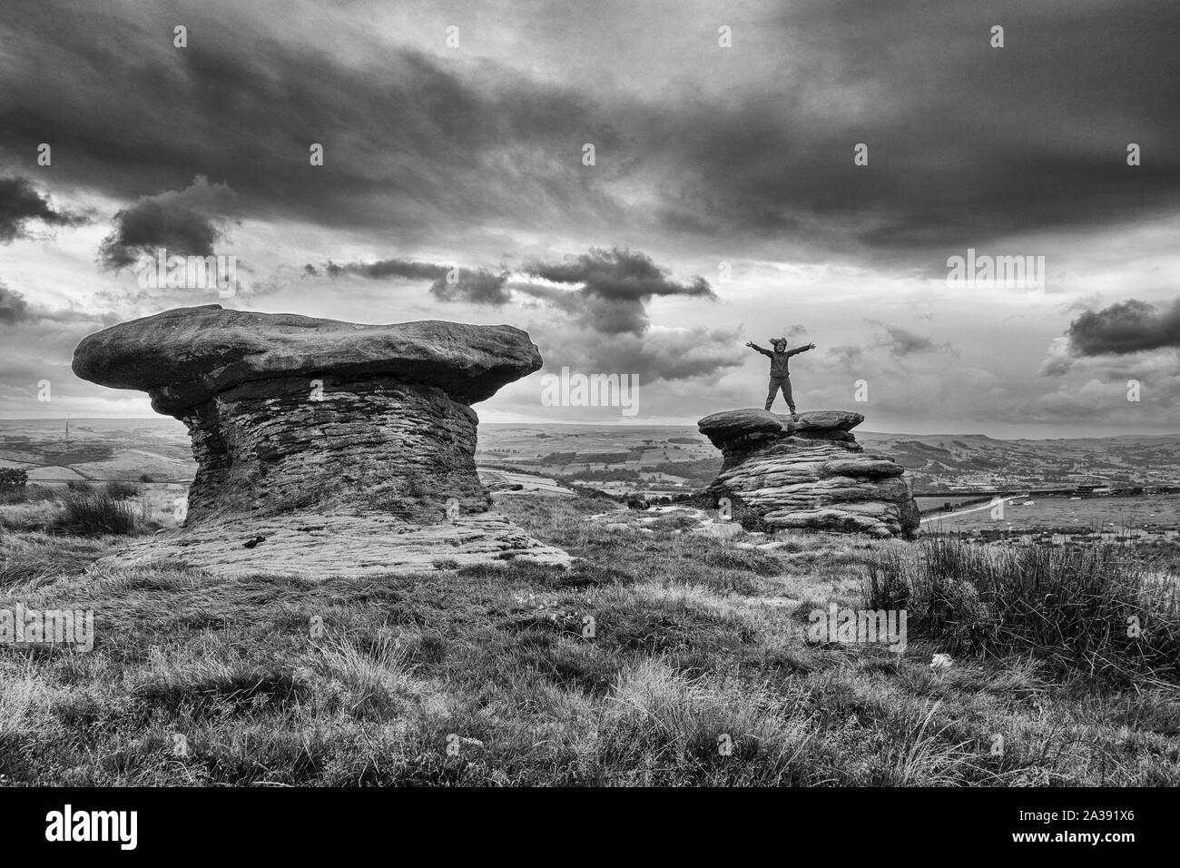 Person standing on top of the Doubler Stones with arms outstretched, Ilkley Moor, UK Their unusual shape is caused by the top being made of hard grits Stock Photo