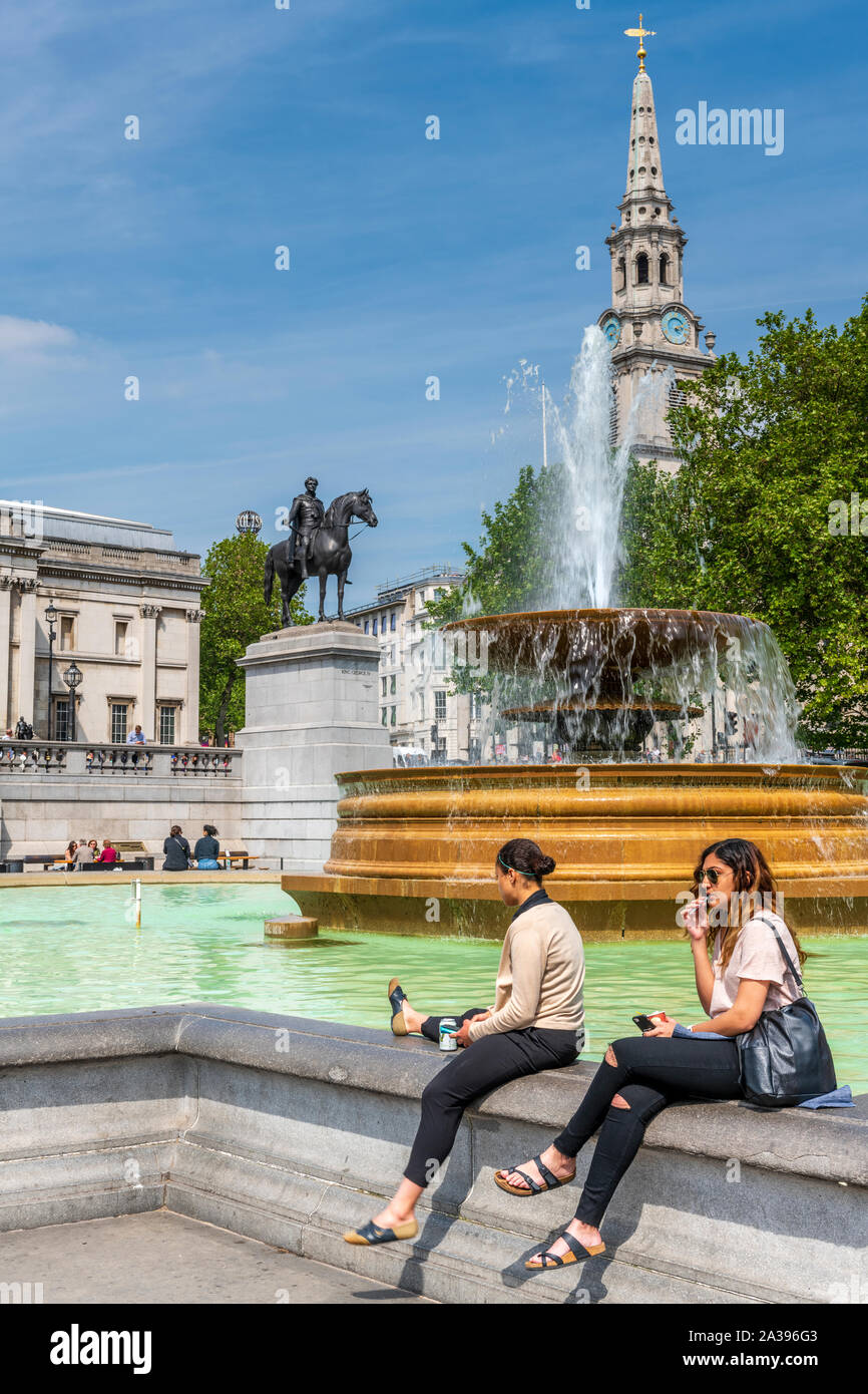 Two young girls enjoy the sunshine sitting on the edge of a fountain in Trafalgar Square, central London. Stock Photo