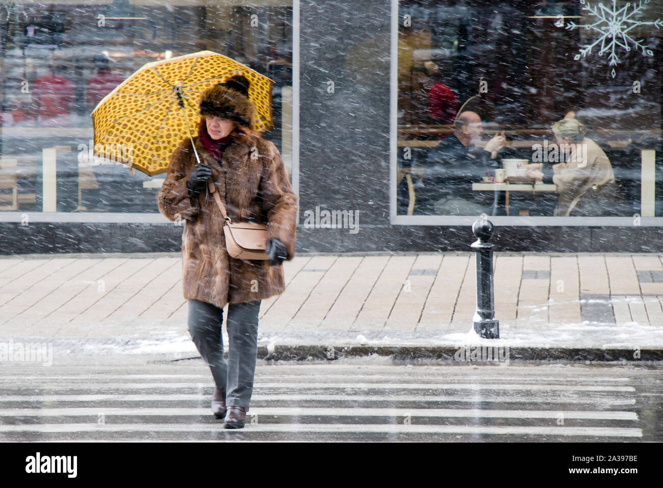 Belgrade, Serbia - January 3, 2019: One mature women in furs crossing the street  under umbrella on snowy day, and elder couple dining together behind Stock Photo