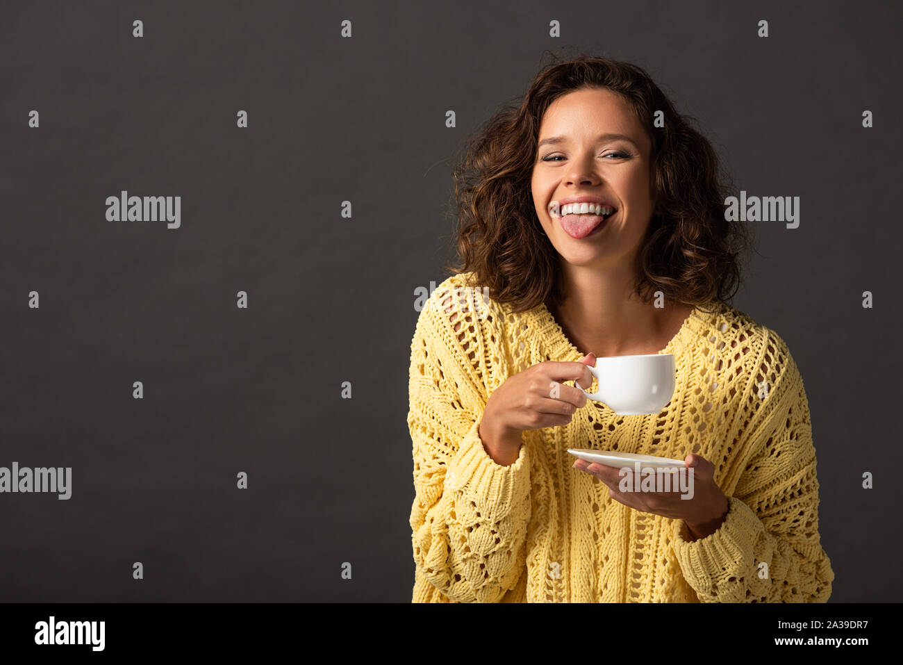 happy curly woman in yellow knitted sweater showing tongue and holding cup of coffee and saucer on black background Stock Photo