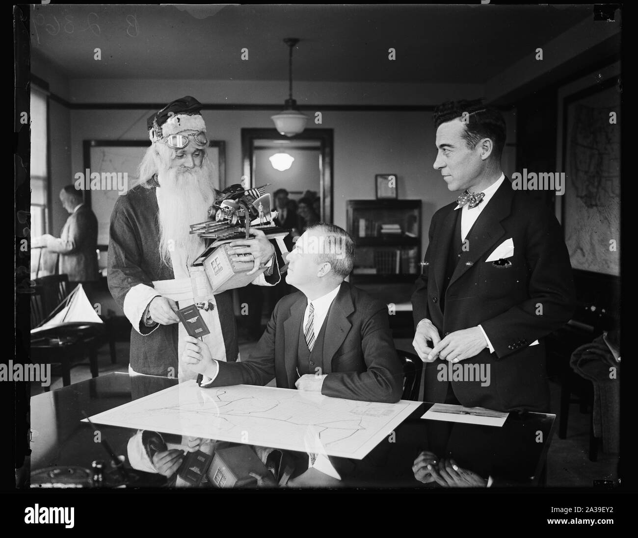 Santa Claus receives aeroplane pilot's license from Assistant Secretary of Commerce. Although there may not be sufficient snow for his reindeer sleigh, Santa Claus will still be able to deliver his load of presents on time this Christmas by using the air route. The old saint called at the Commerce Department in Washington today where he is shown receiving an aeroplane pilot's license from Assistant Secretary of Commerce. for Aeronautics William P. MacCracken, while Clarence M. Young (right) Director of Aeronautics, Department of Commerce, looks on. Airway maps and the assurance that the lights Stock Photo