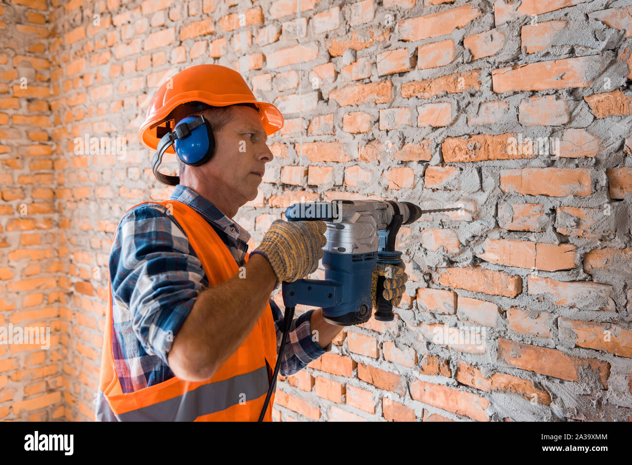 mature builder in helmet using hammer drill near brick wall Stock Photo