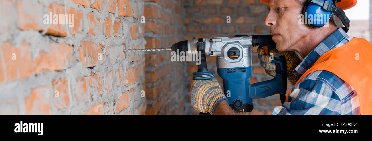 panoramic shot of mature builder using hammer drill near brick wall Stock Photo