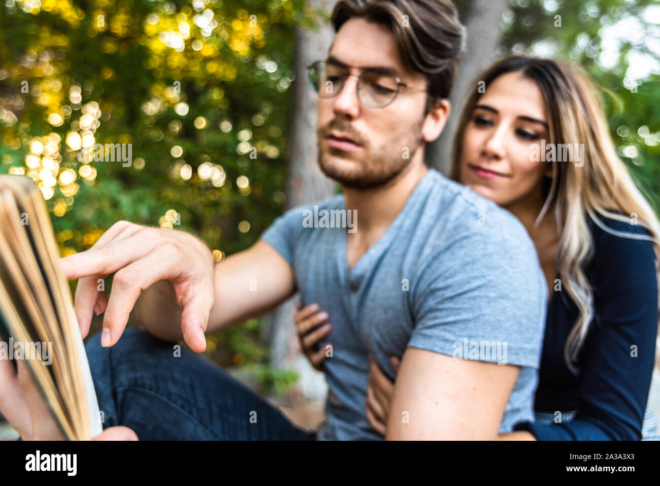 A man's hand pointing to a book, with couple embraced in the background out of focus, in a park. Stock Photo