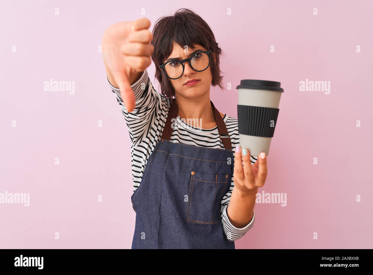 Young beautiful barista woman wearing glasses holding coffee over isolated pink background with angry face, negative sign showing dislike with thumbs Stock Photo