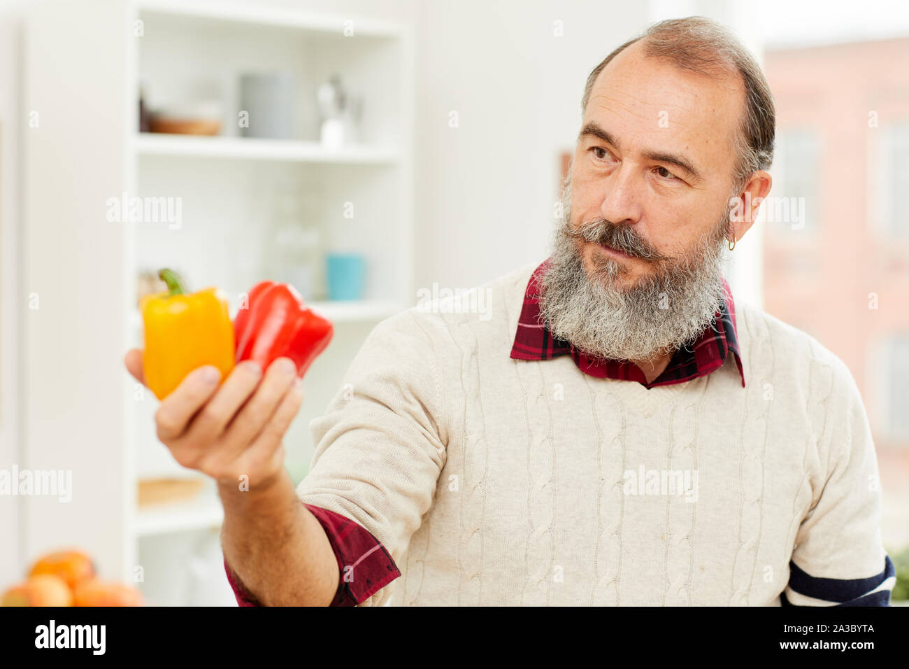 Portrait of bearded senior man holding fresh bell peppers while cooking vegetables in kitchen, copy space Stock Photo