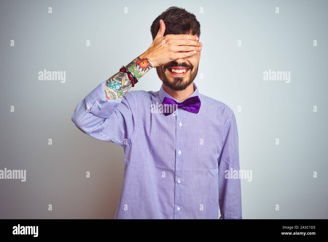 Young man with tattoo wearing purple shirt and bow tie over isolated white background smiling and laughing with hand on face covering eyes for surpris Stock Photo - Alamy