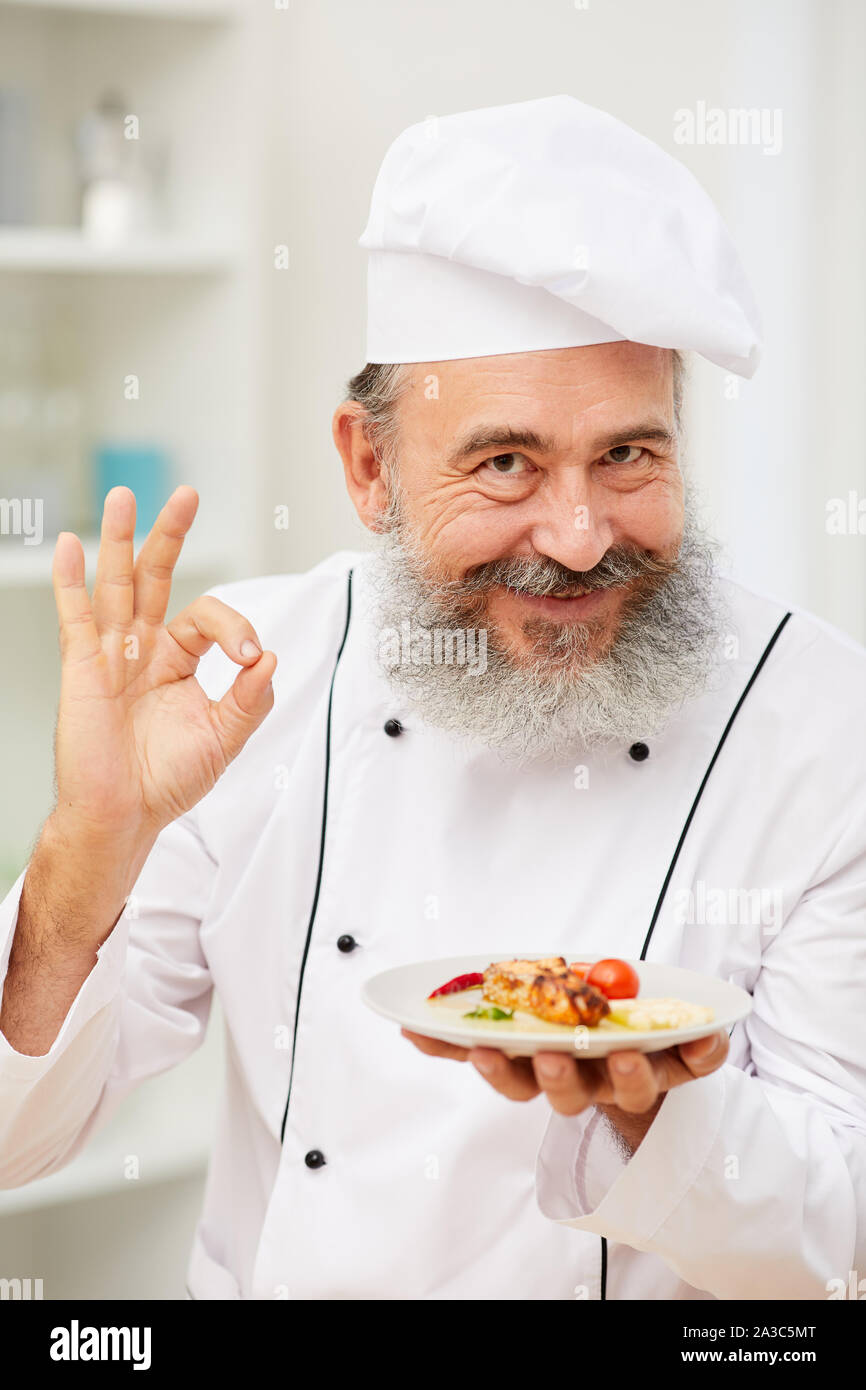 Portrait of cheerful senior chef presenting beautiful Italian dish and showing OK sign while posing in restaurant kitchen Stock Photo