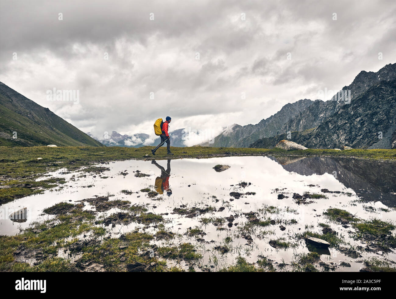 Tourist with yellow backpack is walking near lake with his reflection at green valley in Karakol national park, Kyrgyzstan Stock Photo