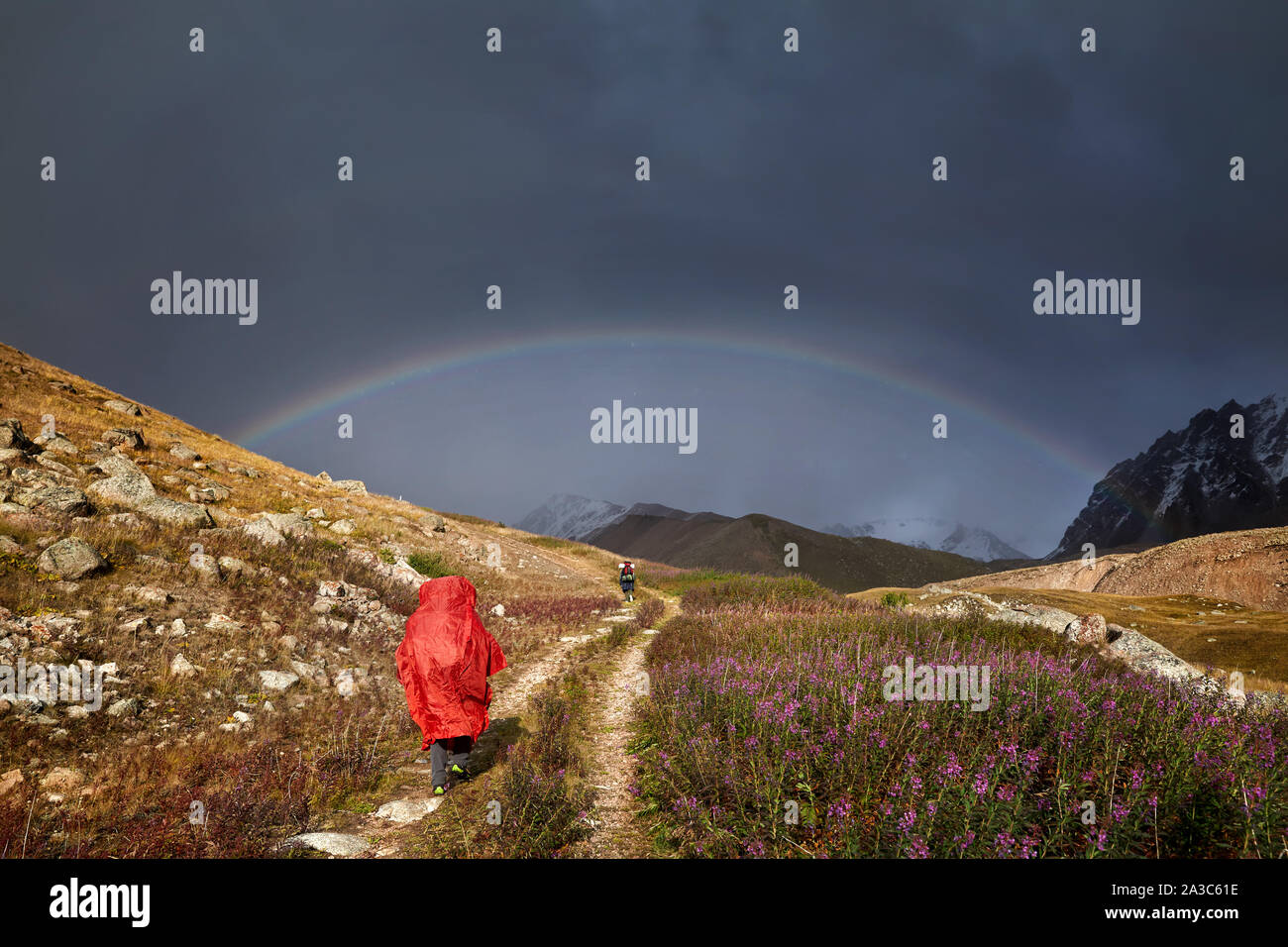 Tourist in the red rain cover is walking down the mountain road with rainbow at background Stock Photo