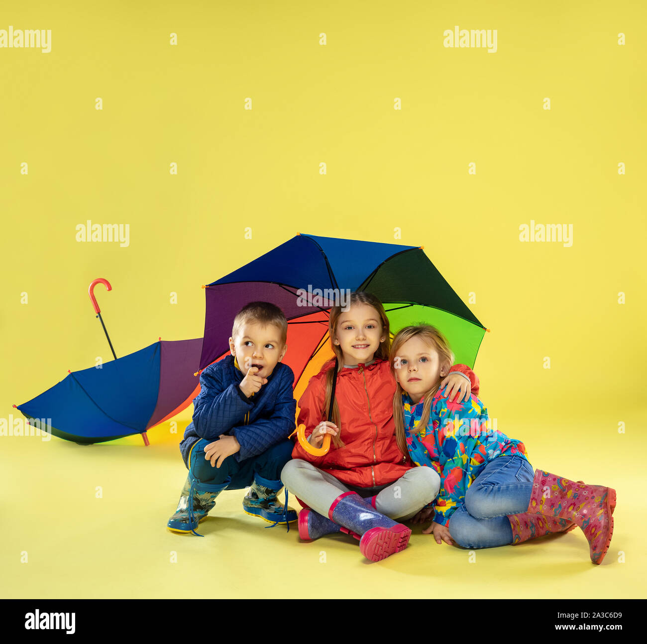 A full length portrait of a bright fashionable kids in a raincoat and  umbrella of rainbow colors on yellow studio background. Autumn and spring  fashion for kids. Cute stylish girls and boy.
