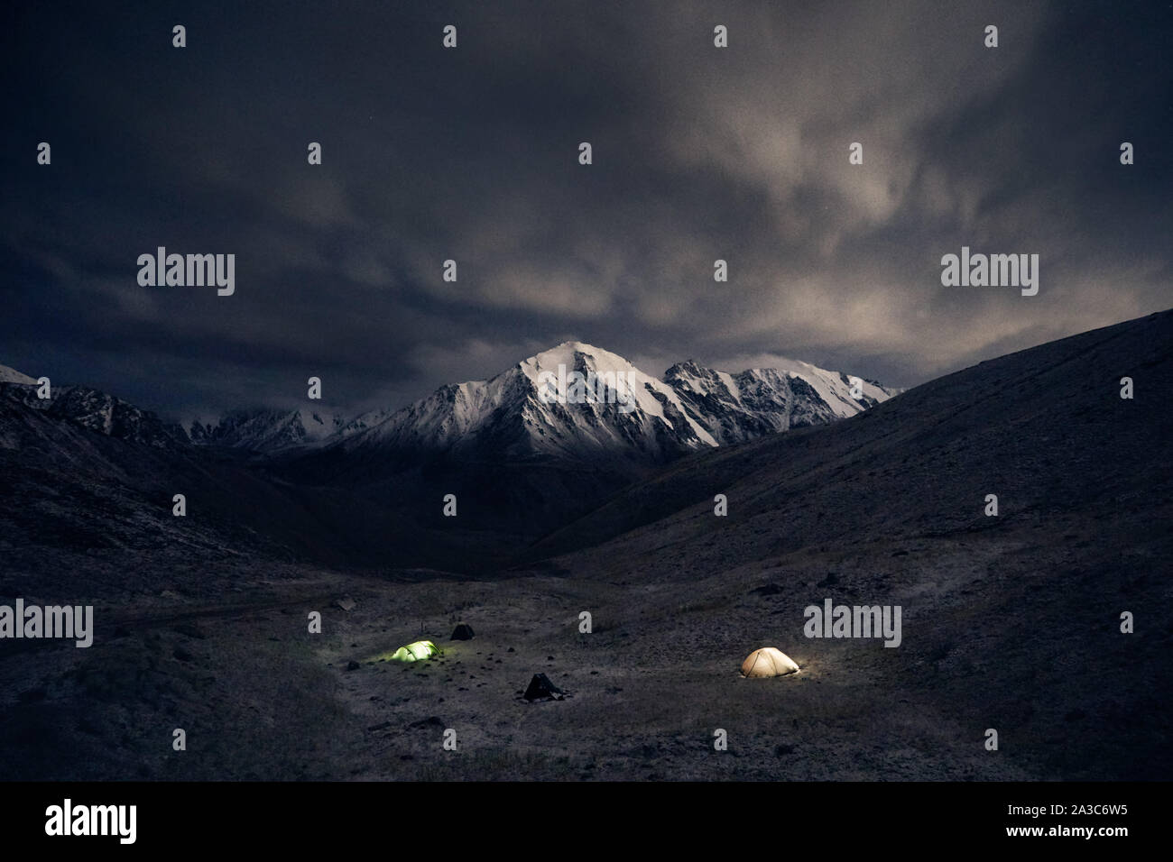 Dramatic landscape of mountain range at night with camping from tents with light at dark cloudy sky background Stock Photo