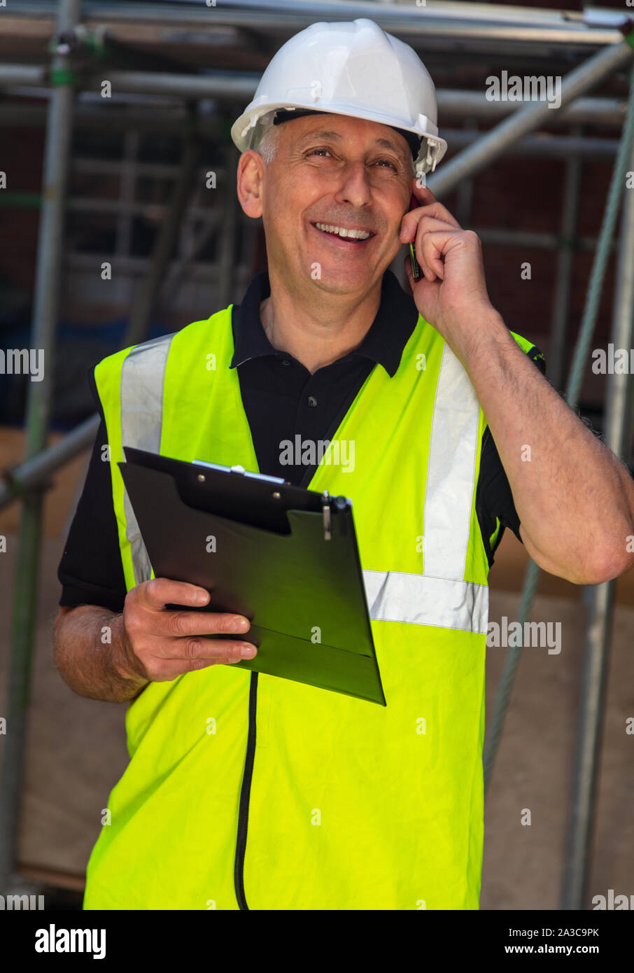 Male builder foreman, worker, contractor or architect on construction site holding black clipboard and talking on his cell phone Stock Photo