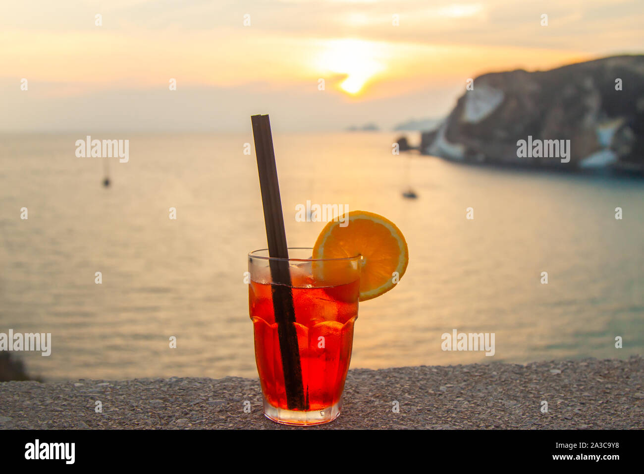 Happy young woman with orange cocktail in glass drink at sunset in front of the sea on Ponza island coast, on a wall with view of the ocean. Smiling Stock Photo
