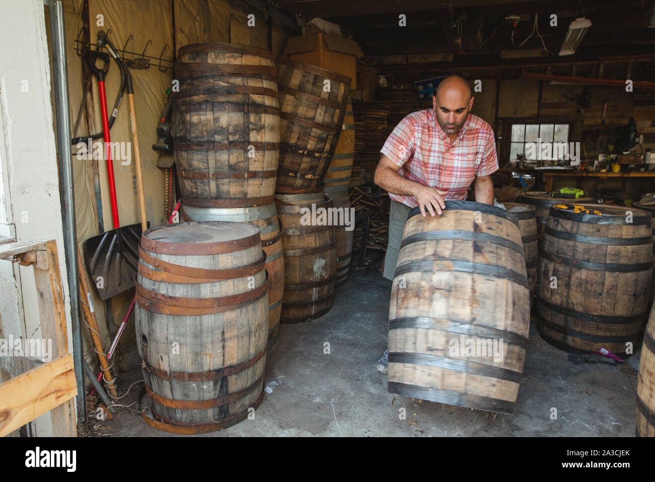 A man wheels a large wood bourbon barrel from a stack in a garage Stock Photo