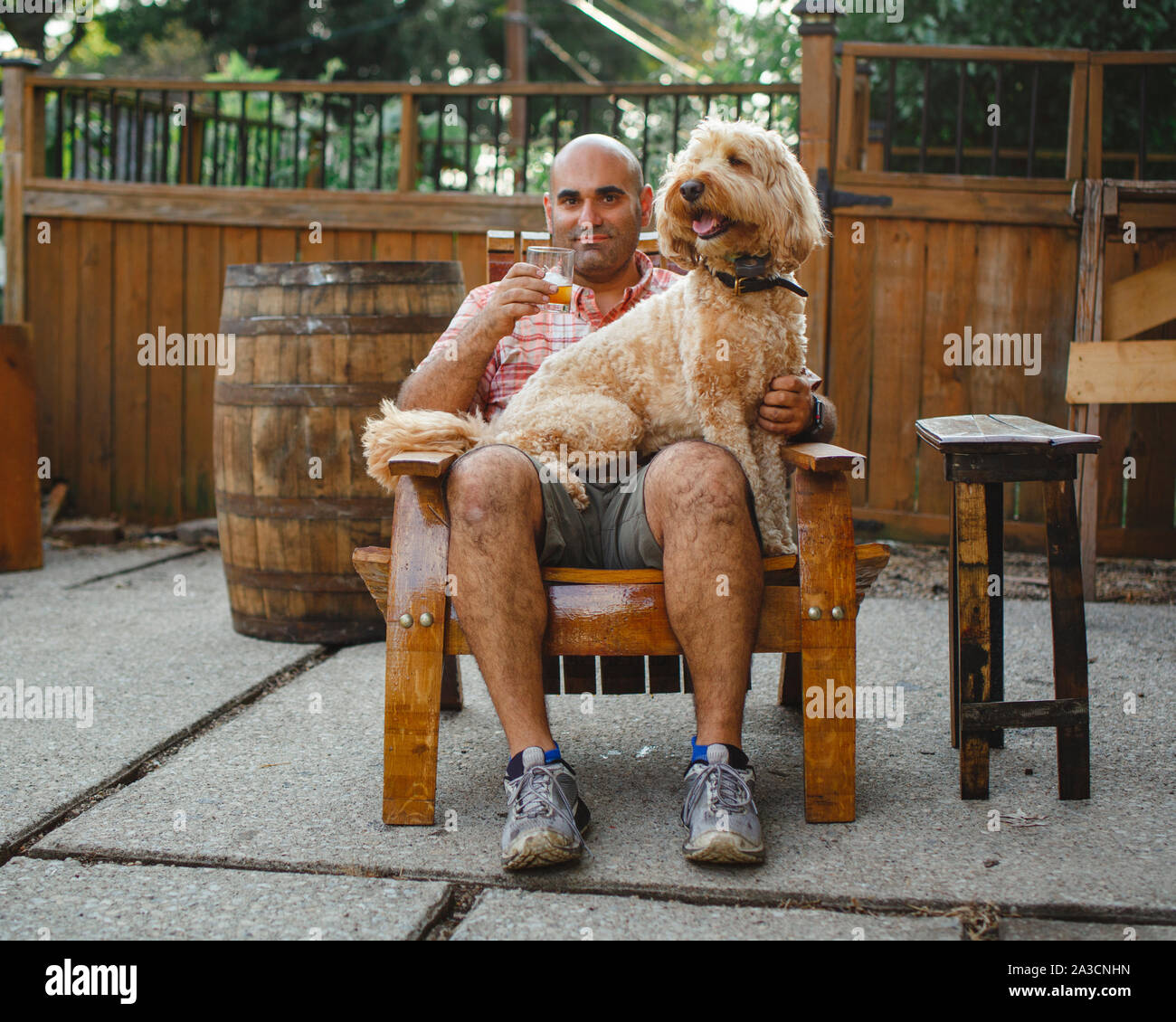A happy man sits with glass of bourbon and dog on lap on wooden chair Stock Photo