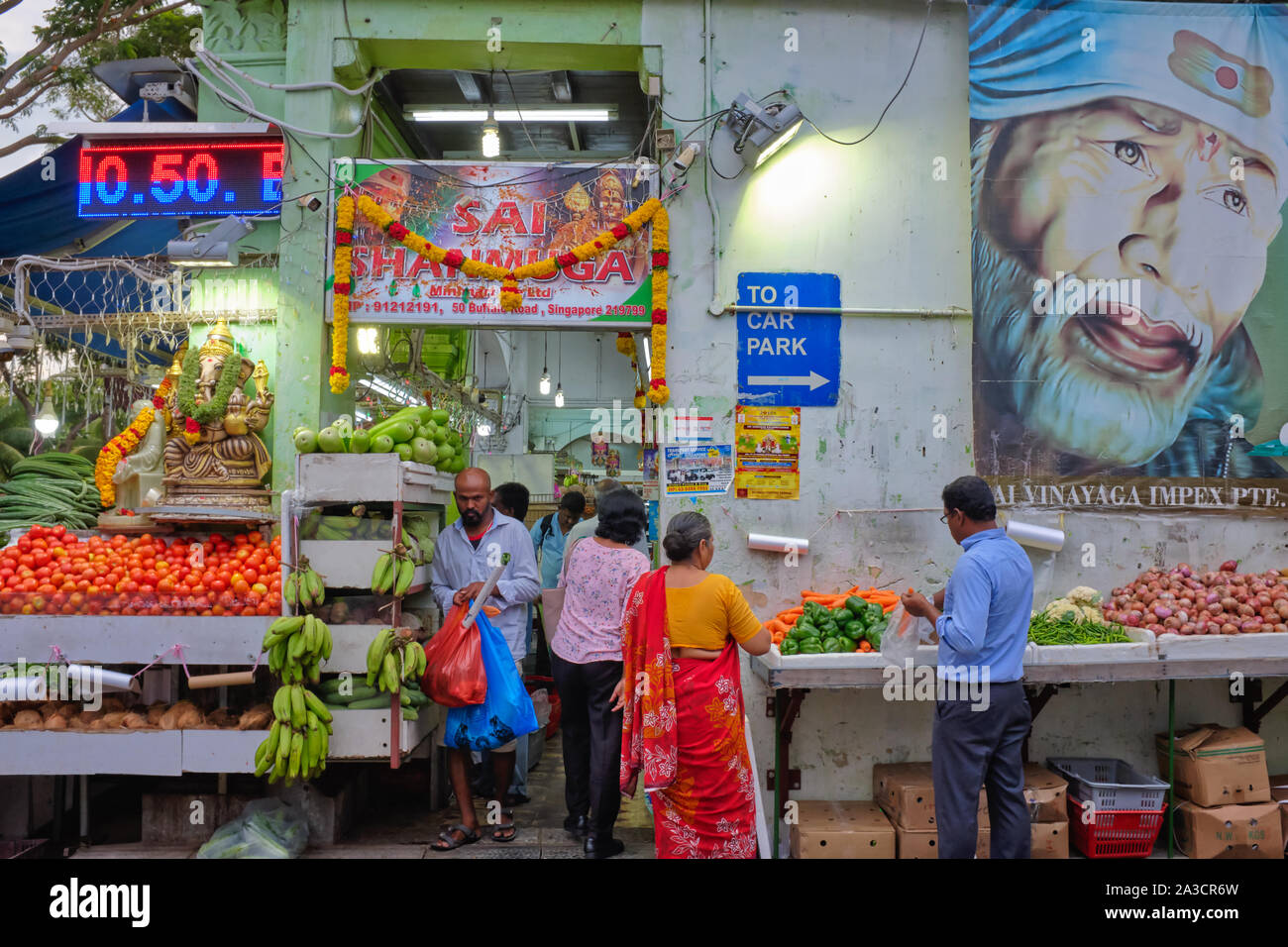 An open-air fruit and vegetable stall overseen by an oversized portrait of Indian saint Sai Baba, and shoppers, in Liittle India area, Singapore Stock Photo